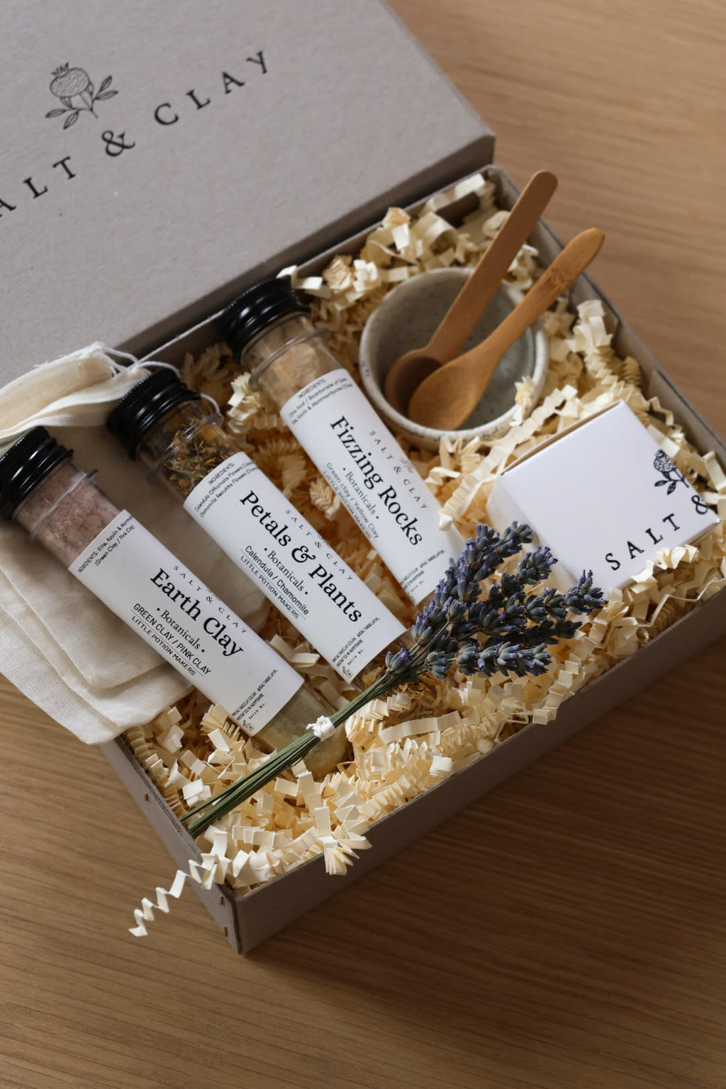A gift box set from Salt & Clay on a wooden table, containing three labeled plastic test tubes of ingredients, a small bowl with wooden spoons, three bath teabags, and dried botanicals, arranged in shredded paper filler.