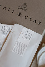 White paper pouch of Salt & Clay clay masks placed on top of a grey gift box with the brand name 'SALT & CLAY' printed on it. A white cotton cloth is partially visible on the right side.