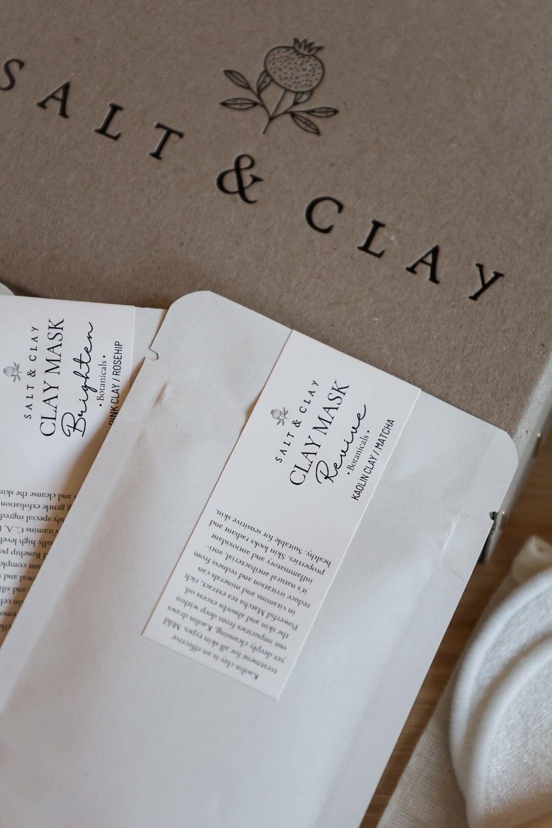 White paper pouch of Salt & Clay clay masks placed on top of a grey gift box with the brand name 'SALT & CLAY' printed on it. A white cotton cloth is partially visible on the right side.