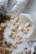 White herbal tea cubes spilling from a small white mesh bag, with dried chamomile flowers and two glass jars on a white surface.