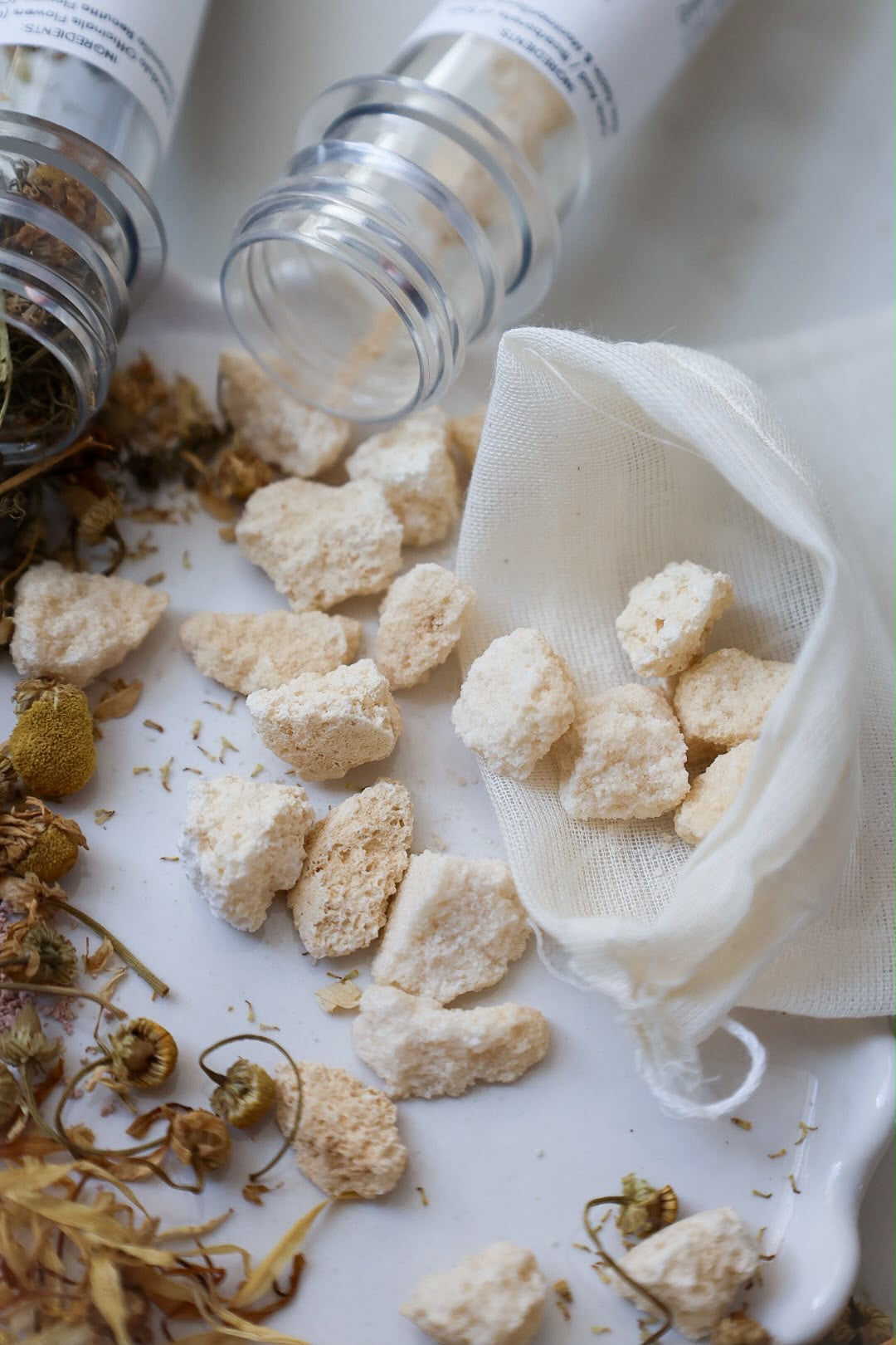 White herbal tea cubes spilling from a small white mesh bag, with dried chamomile flowers and two glass jars on a white surface.