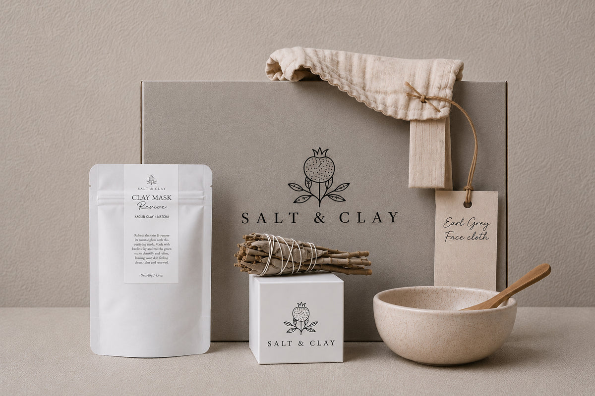 A styled clay face mask gift set from Salt & Clay on a neutral beige surface against a textured beige wall. The set includes a large gray box with the 'SALT & CLAY' logo, a white pouch of clay face mask, a bath bomb, a sage smudge wand, a beige linen face cloth, and a beige plastic bowl with a bamboo spoon.