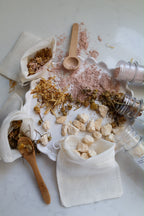 A styled flat-lay of a natural bath tea bathtime play set on a white marble surface, featuring white mesh drawstring bags filled with dried botanicals, loose pink clay, pale yellow fizzing bath rocks, wooden spoons, and clear plastic test tubes.
