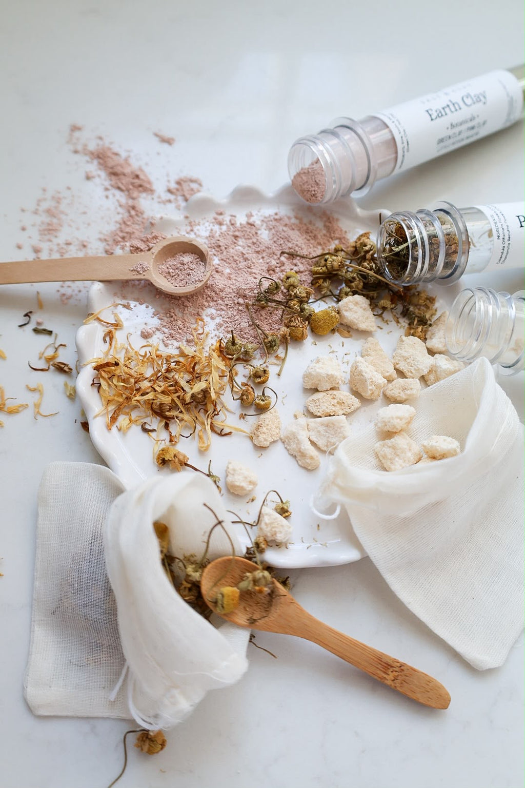An overhead styled flat-lay of a DIY earth clay bath time play set for children. The scene features several clear plastic test tubes with white labels reading 'Earth Clay', some open, showing pink clay and dried botanicals of chamomile and calendula petals.