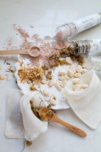 An overhead styled flat-lay of a DIY earth clay bath time play set for children. The scene features several clear plastic test tubes with white labels reading 'Earth Clay', some open, showing pink clay and dried botanicals of chamomile and calendula petals.