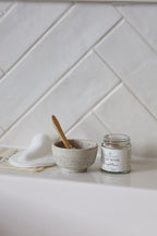 A small speckled ceramic bowl with a wooden spoon sits on a white countertop beside a white folded towel and a clear glass jar labeled 'CLAY FACE MASK'. The background is a white herringbone tile wall.