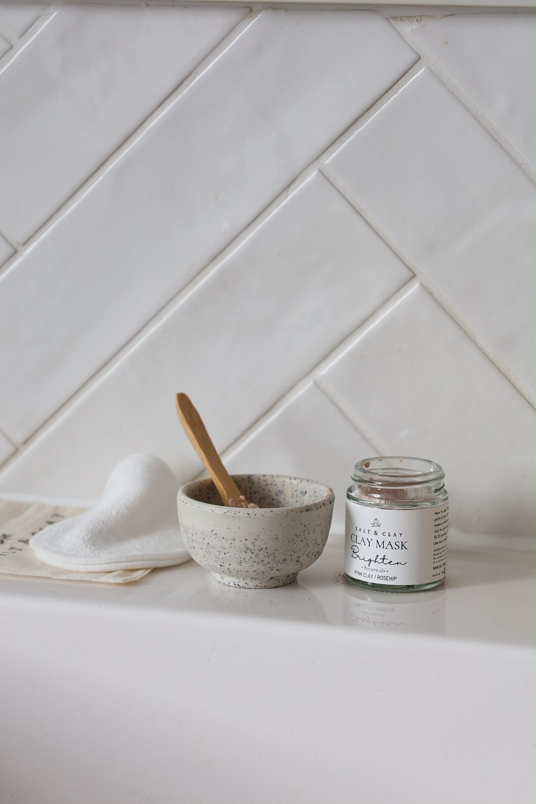 A small speckled ceramic bowl with a wooden spoon sits on a white countertop beside a white folded towel and a clear glass jar labeled 'CLAY FACE MASK'. The background is a white herringbone tile wall.