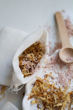 A white fabric cotton pouch filled with dried botanicals and pink powdered clay, styled on a white surface with scattered loose pink clay, dried Calendula petals, and a wooden spoon.