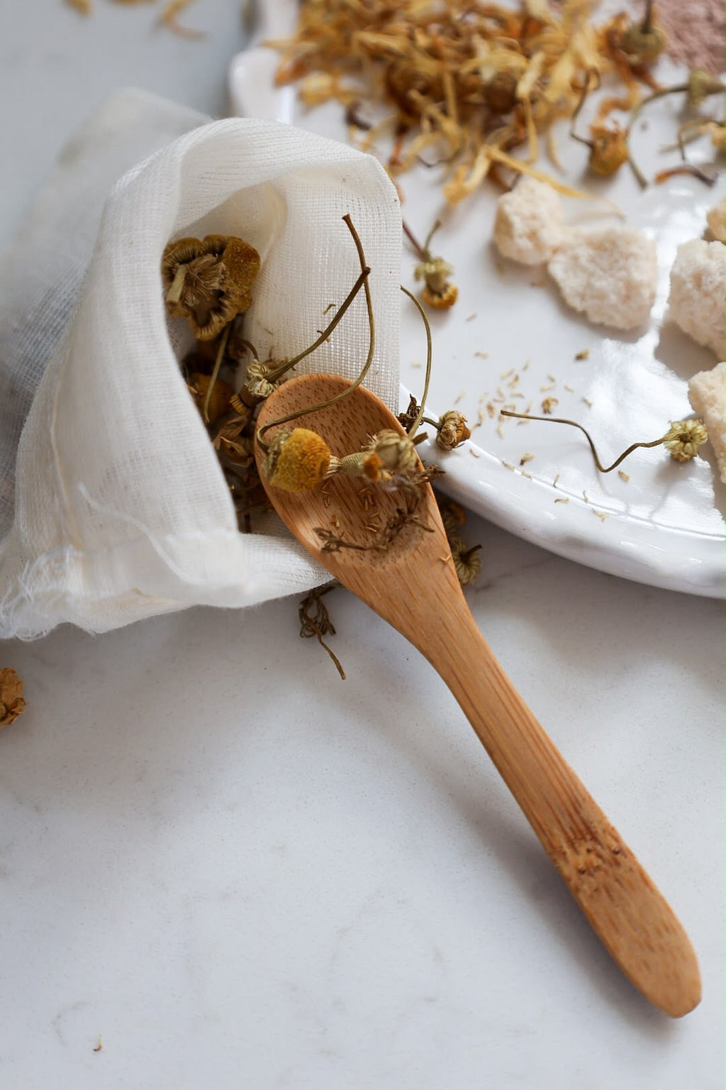 A wooden spoon filled with dried chamomile flowers spilling out of a white gauze bag onto a white marble surface, with a white ceramic dish in the background.