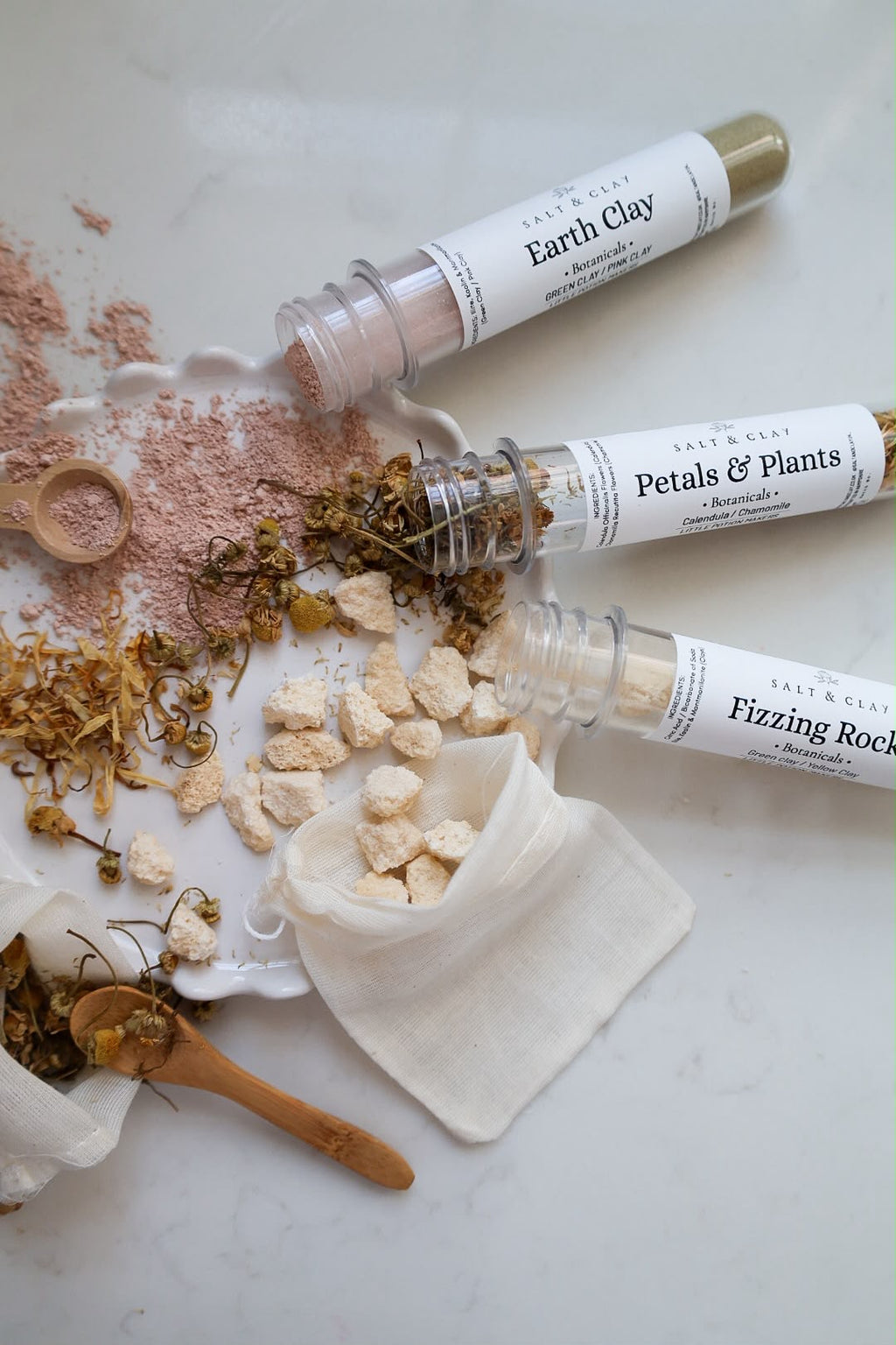 Three clear plastic test tubes of natural clay labeled 'Earth Clay', 'Petals & Plants', and 'Fizzing Rocks' are arranged on a white marble surface with the powders and chunks spilled out, alongside a small white cloth pouch, wooden spoon, and dried botanicals.