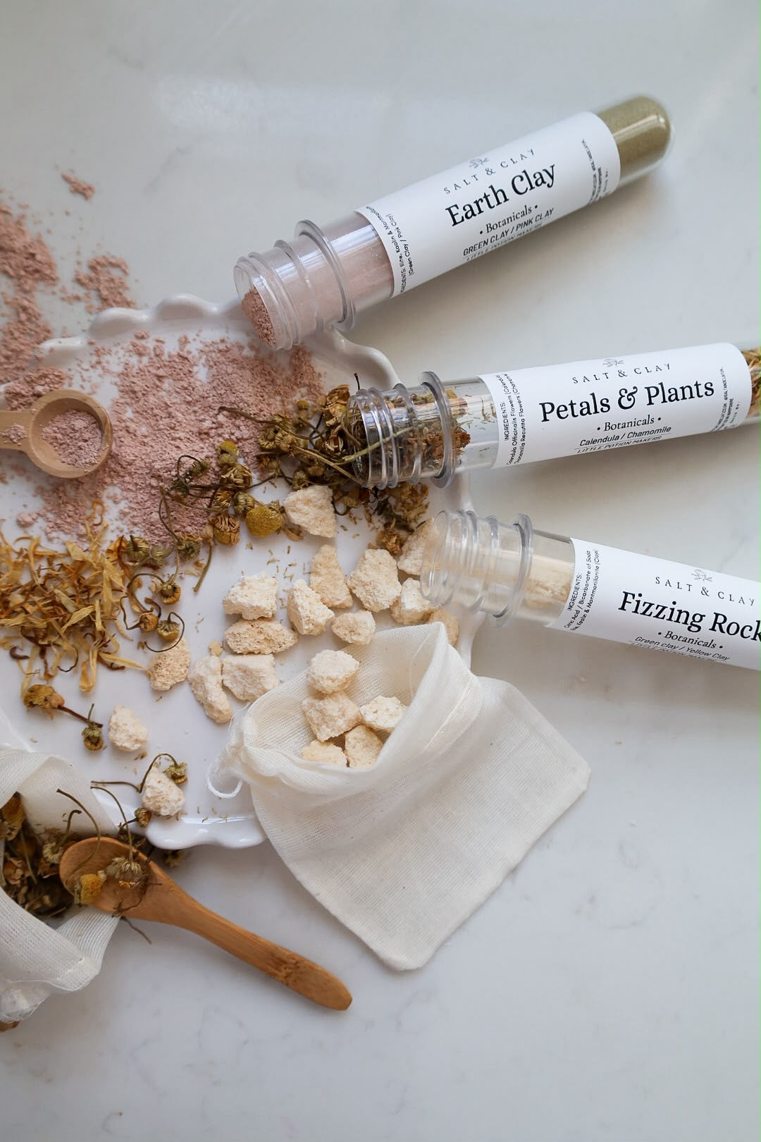 Three clear plastic test tubes of natural clay labeled 'Earth Clay', 'Petals & Plants', and 'Fizzing Rocks' are arranged on a white marble surface with the powders and chunks spilled out, alongside a small white cloth pouch, wooden spoon, and dried botanicals.