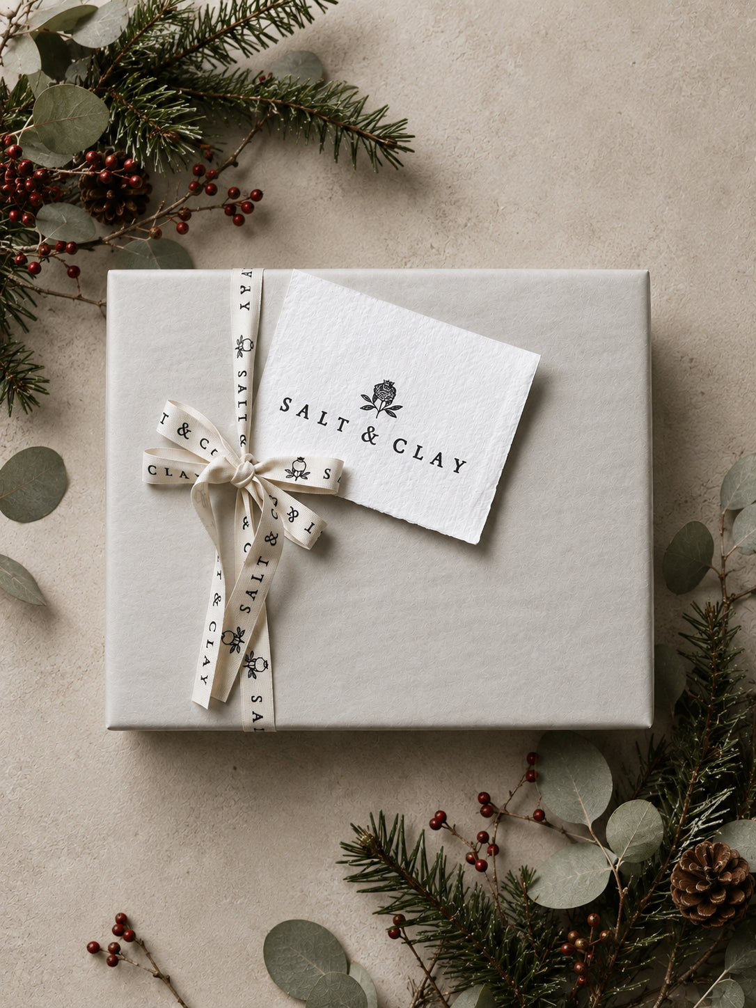 A square gift box wrapped in light beige paper, tied with a cream ribbon printed with the brand name 'SALT & CLAY', and topped with a matching branded paper tag. The box is styled on a neutral beige surface with eucalyptus leaves, pine branches, and red berries around it.