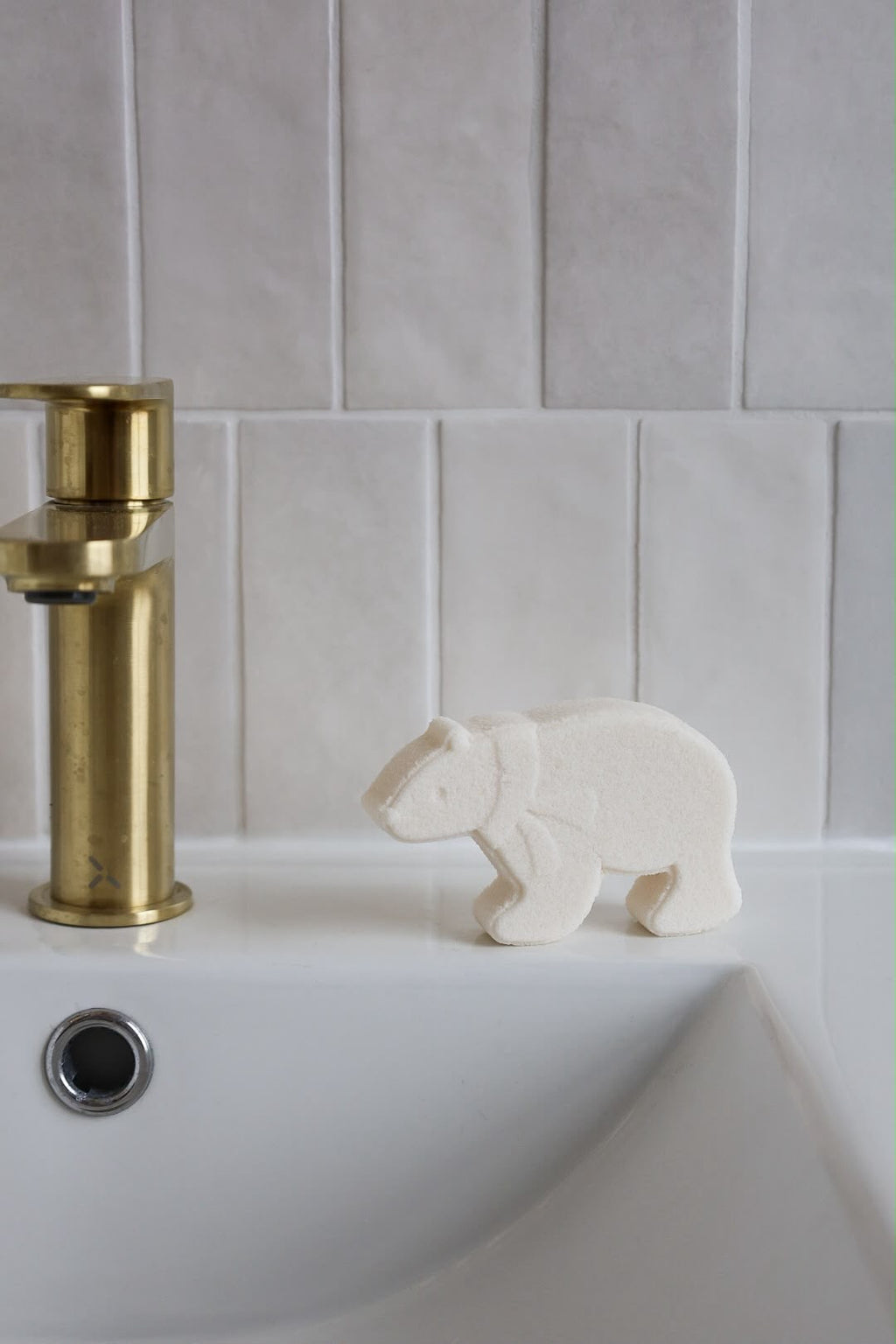 A small white bear-shaped fizzing bath bomb sits on the edge of a white sink next to a brushed gold faucet, set against a light gray tiled wall.