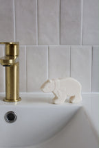A small white bear-shaped fizzing bath bomb sits on the edge of a white sink next to a brushed gold faucet, set against a light gray tiled wall.