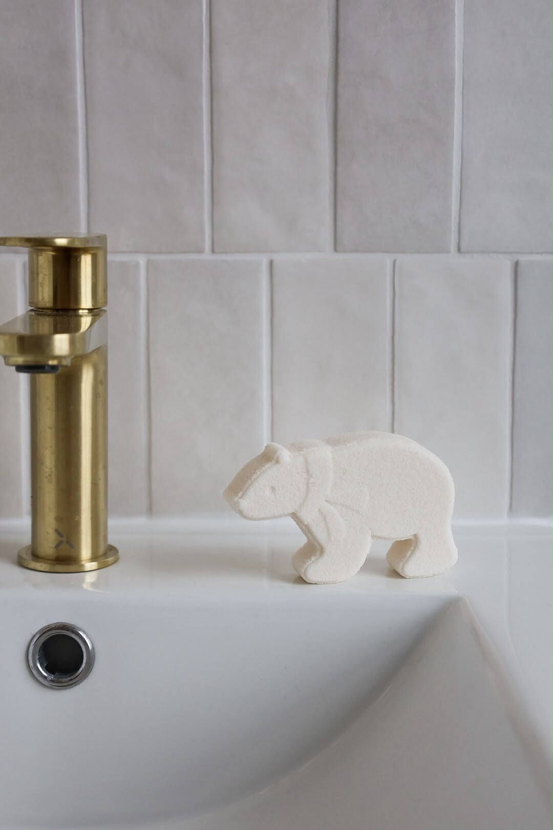 A small white bear-shaped fizzing bath bomb sits on the edge of a white sink next to a brushed gold faucet, set against a light gray tiled wall.