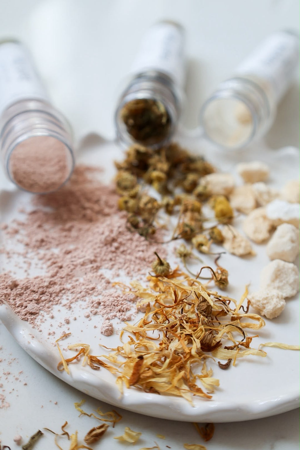 A close-up, artistic flat-lay of a natural bath tea blend on a white ceramic plate. The scene features loose dried yellow Calendula flowers, light brown herbal Chamomile, and a fine dusty pink clay powder spilling from three clear plastic test tubes lying on their sides.
