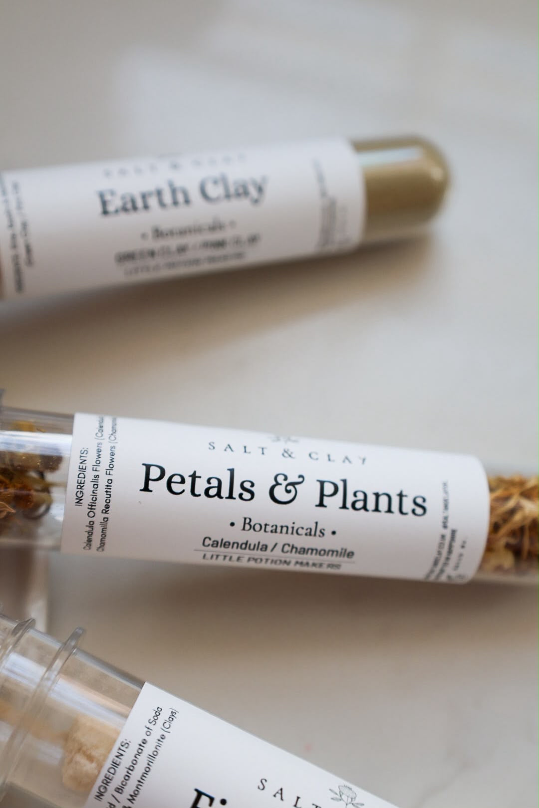 Three clear plastic tubes containing herbal salts or powders, each with a white paper label and a black cap, arranged diagonally on a light beige surface. The labels read 'Salt & Clay', 'Earth Clay', 'Petals & Plants'.