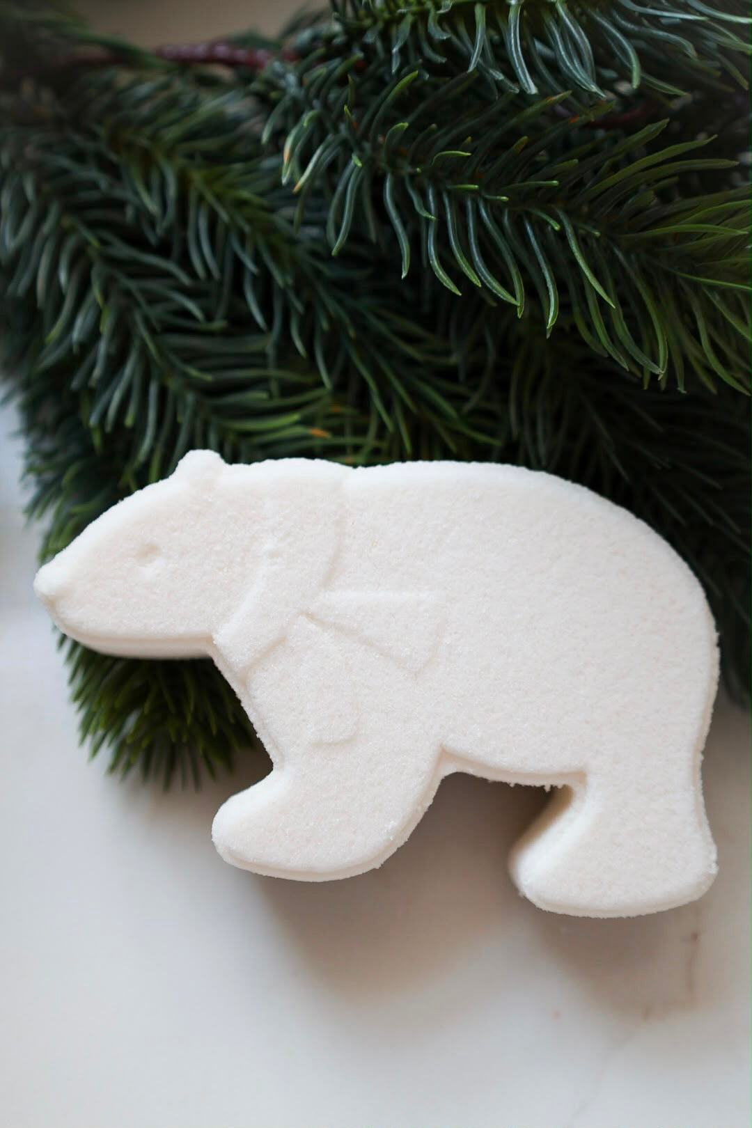 A white polar bear-shaped bath bomb with a small scarf detail, styled on a light surface with green pine branches in the background.