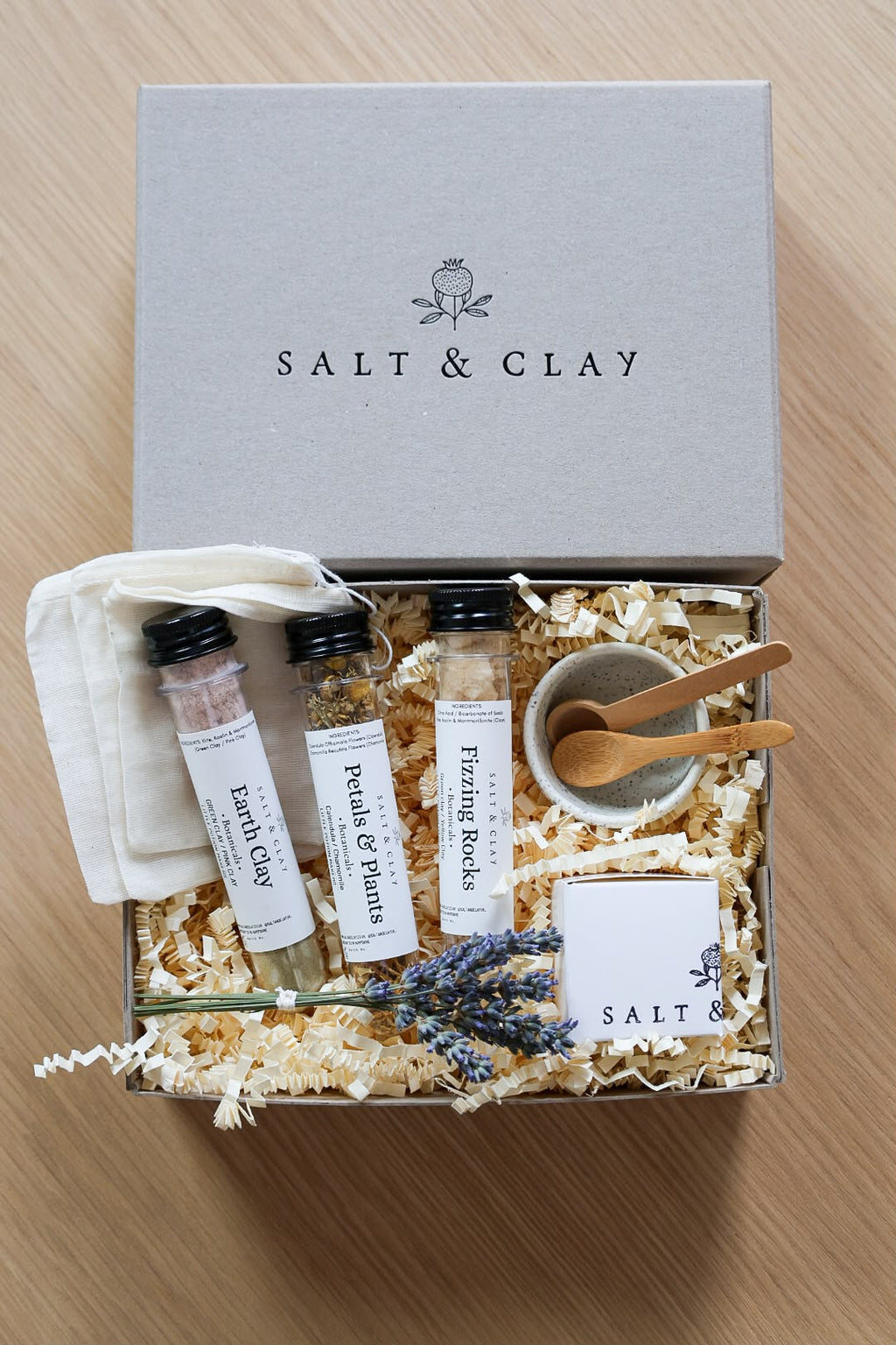 A Salt & Clay gift set arranged in an open box with shredded paper filler on a light wood surface. The set includes a gray branded box lid, three labeled plastic tubes, a small white cloth pouch, a small beige bowl with wooden spoons, lavender sprigs, and a branded card.