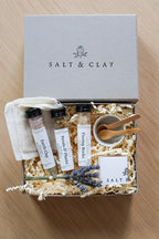 A Salt & Clay gift set arranged in an open box with shredded paper filler on a light wood surface. The set includes a gray branded box lid, three labeled plastic tubes, a small white cloth pouch, a small beige bowl with wooden spoons, lavender sprigs, and a branded card.