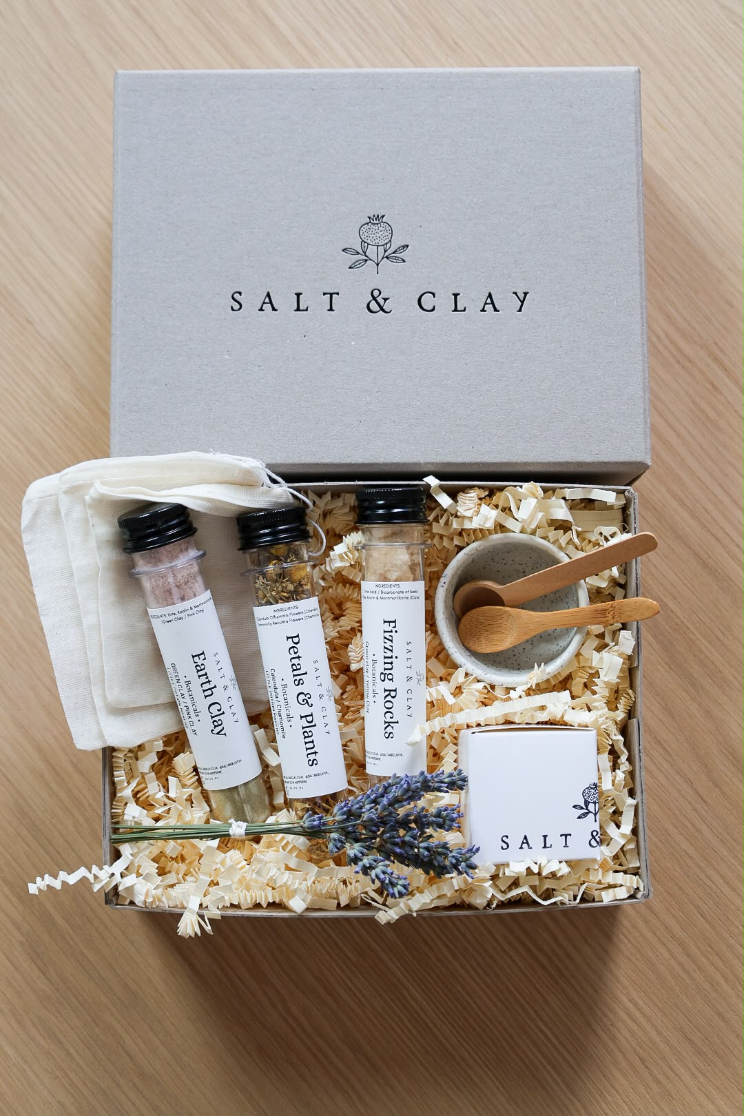 A Salt & Clay gift set arranged in an open box with shredded paper filler on a light wood surface. The set includes a gray branded box lid, three labeled plastic tubes, a small white cloth pouch, a small beige bowl with wooden spoons, lavender sprigs, and a branded card.