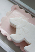 A white, bear-shaped bath bomb resting on a pink scalloped dish, set against a neutral grey background.