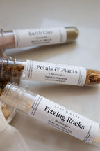 Three clear glass jars labeled 'Salt & Clay' containing different clay mixes: 'Earth Clay', 'Petals & Plants', and 'Fizzling Rocks'. The jars are arranged on a light beige surface with a white cloth in the foreground.